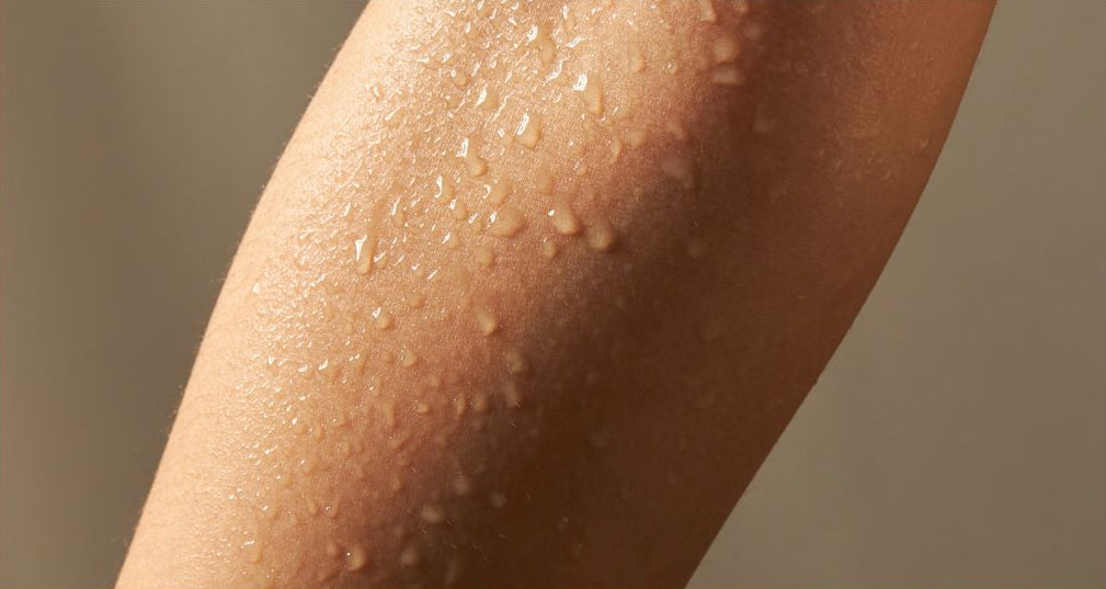 Golden-yellow oil being dropped from glass pipettes onto a textured beige surface, with a close-up of a human arm covered in droplets below.